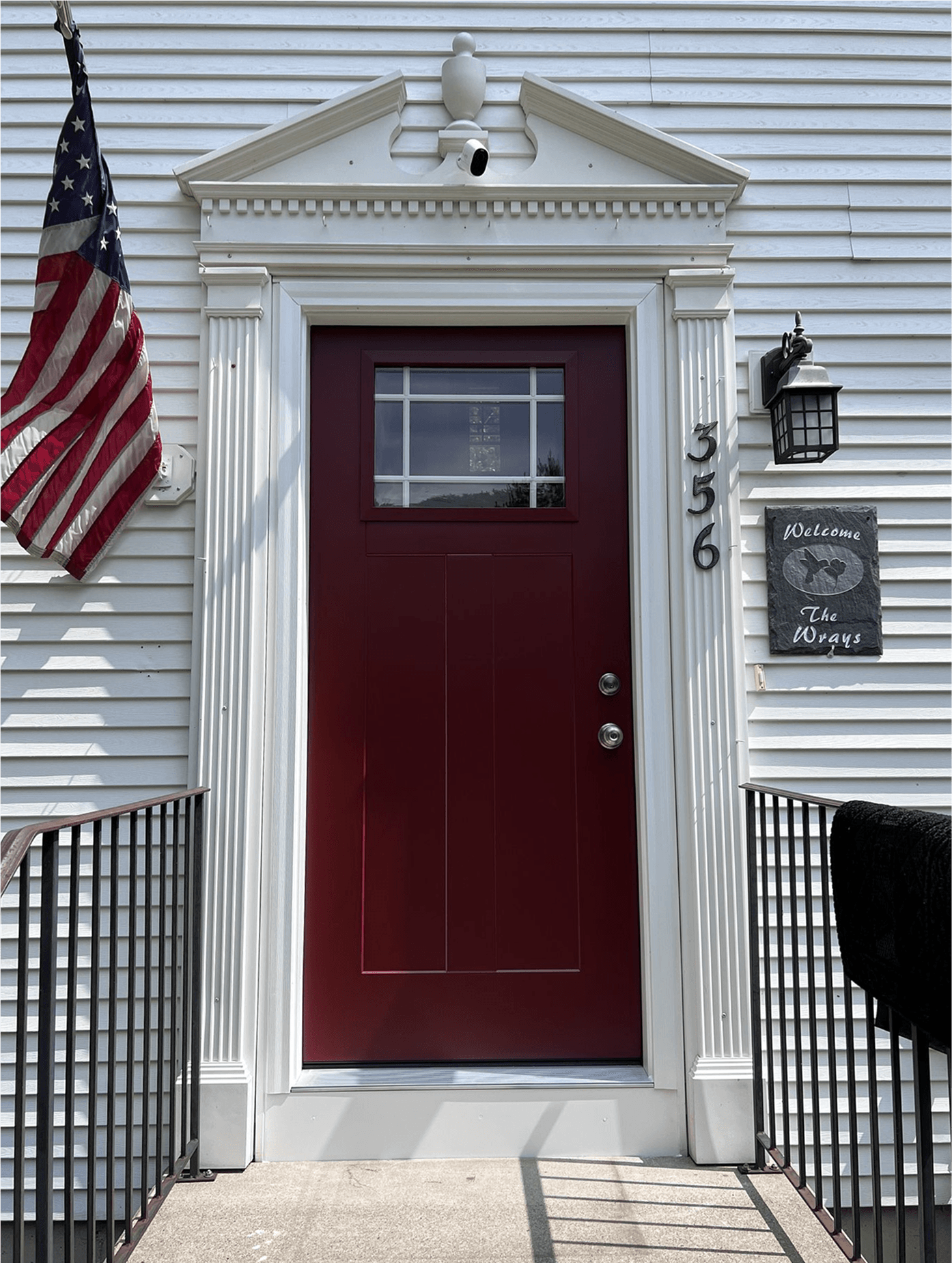 A dark red front door with small window on a white house. An American flag hangs to the left.