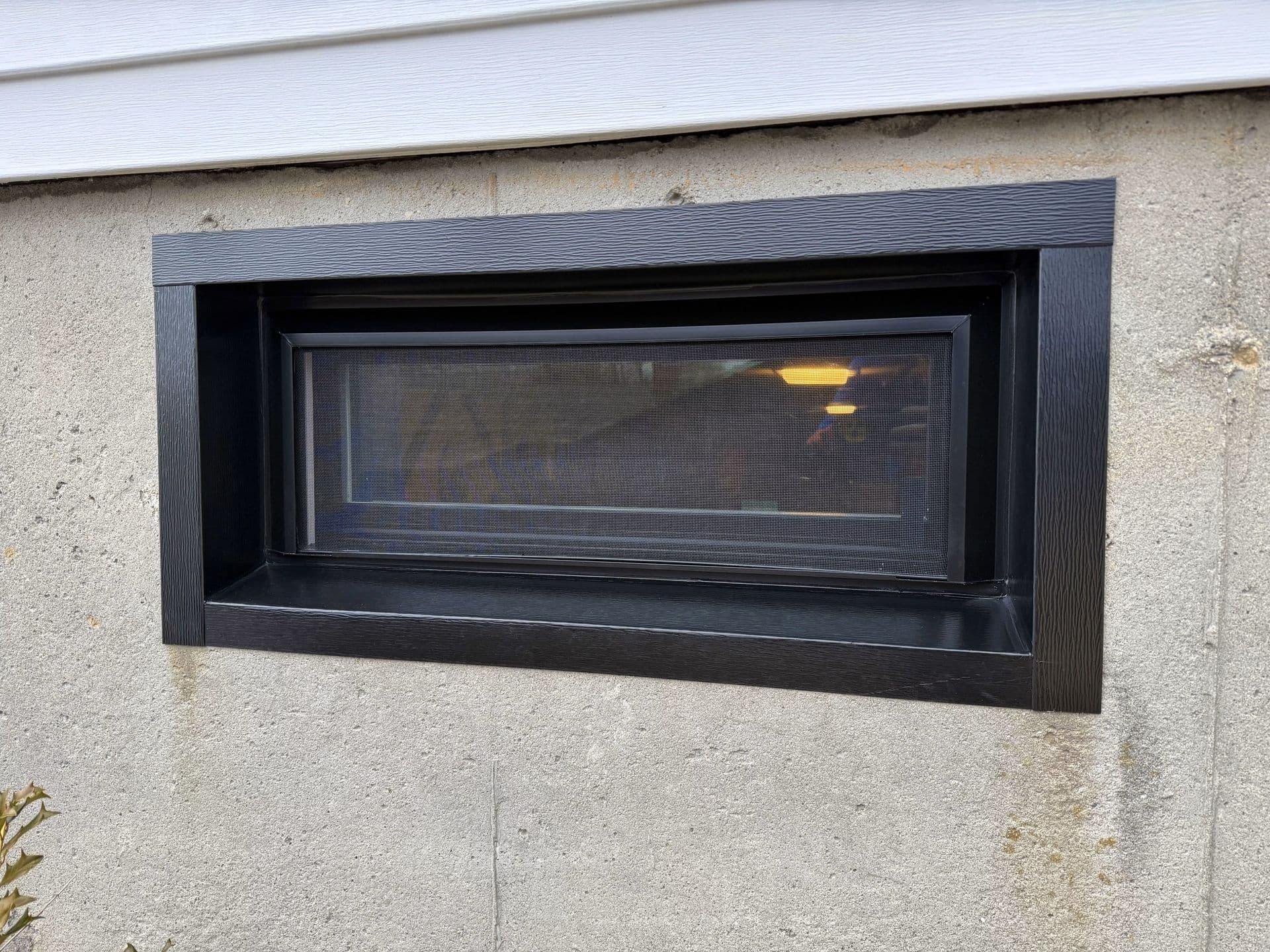 A basement hopper window with black trim from the outside.