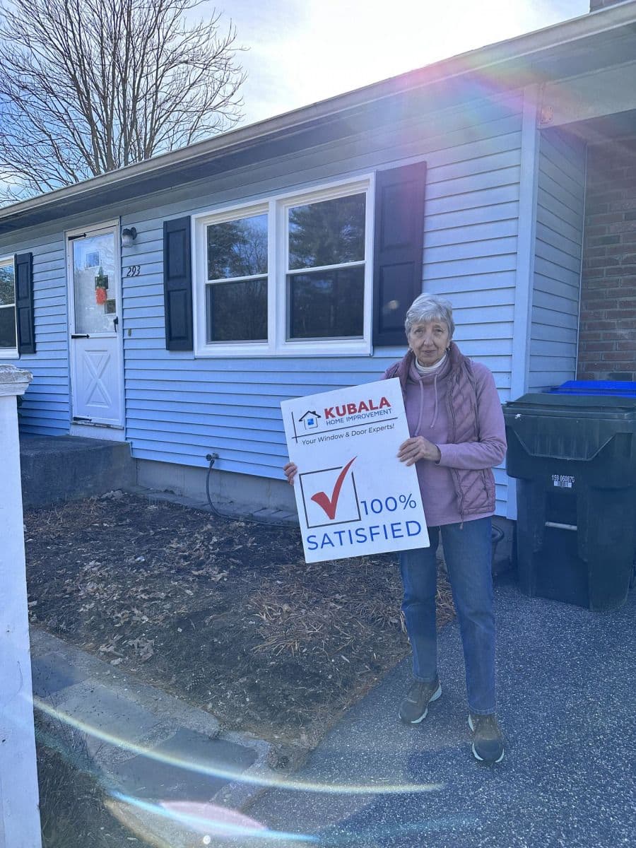 A homeowner holds a sign that reads "100% Satisfied" and stands in front of new replacement windows.
