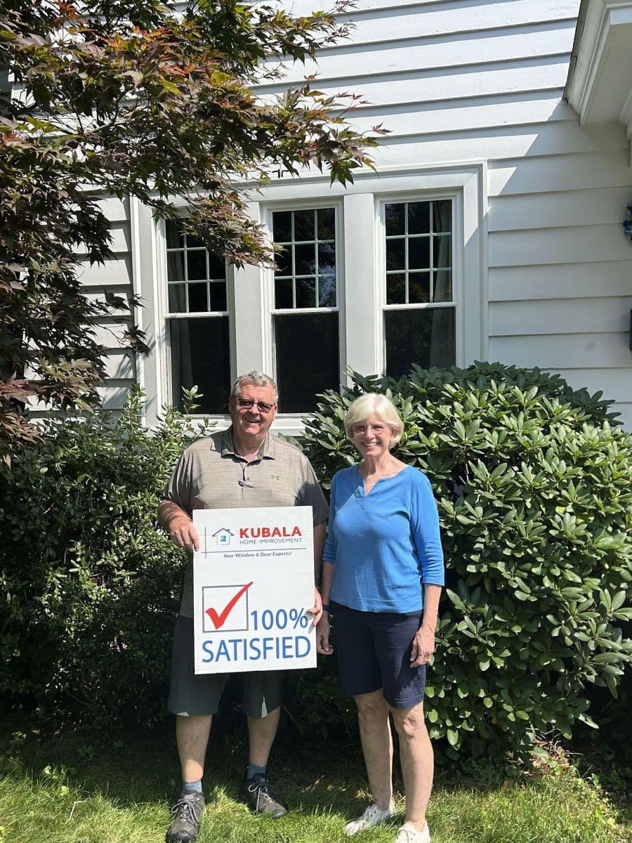 Homeowners hold a sign that reads "100% Satisfied" and stand in front of newly-installed windows.