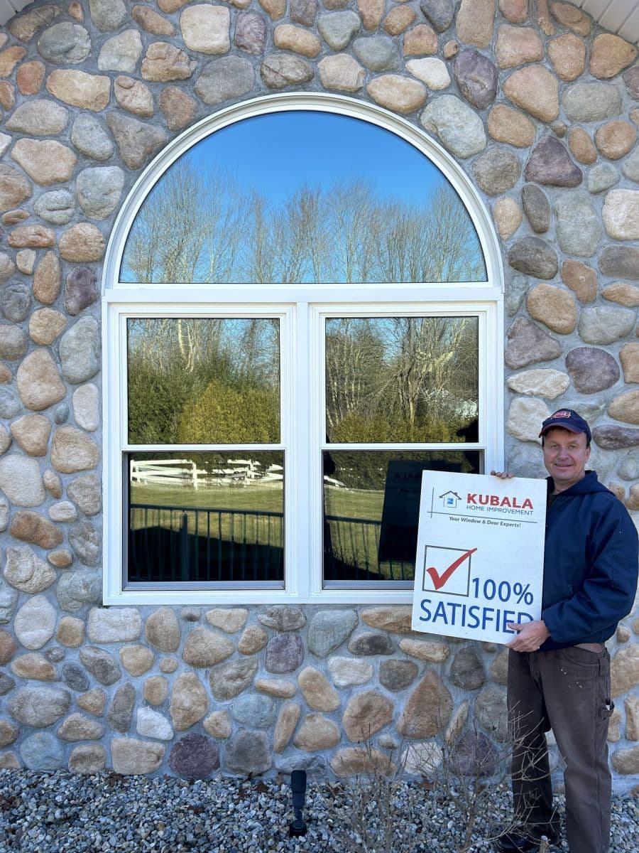 A homeowner holds a sign that reads "100% Satisfied" and stands in front of a new custom replacement window.