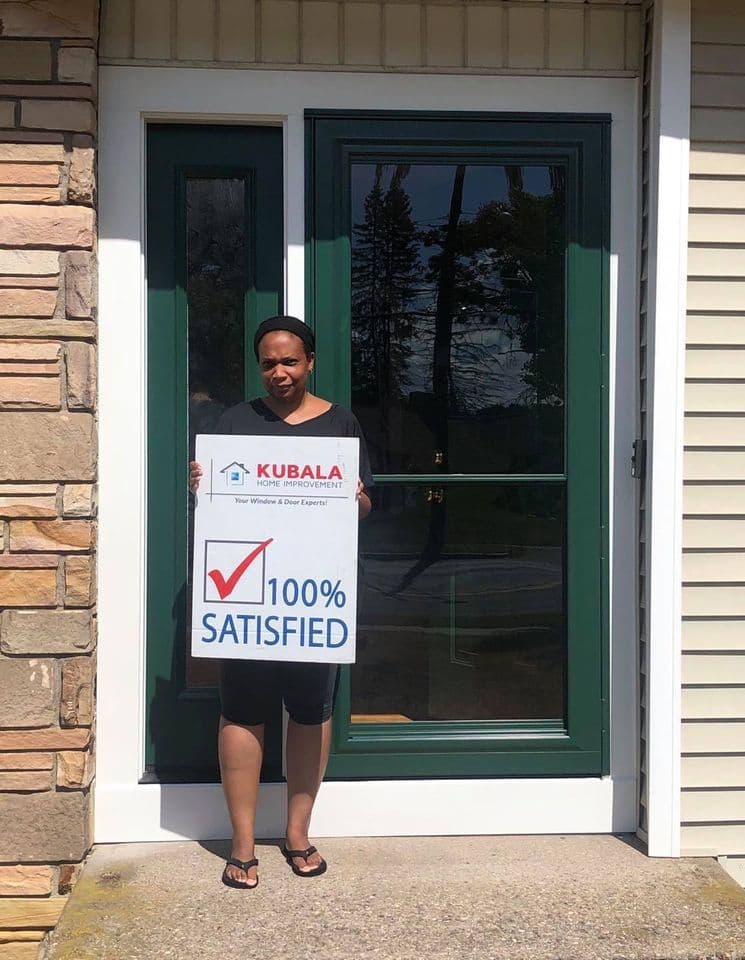 A homeowner holds a sign that reads "100% Satisfied" and stands in front of a new dark green entry door with white trim.