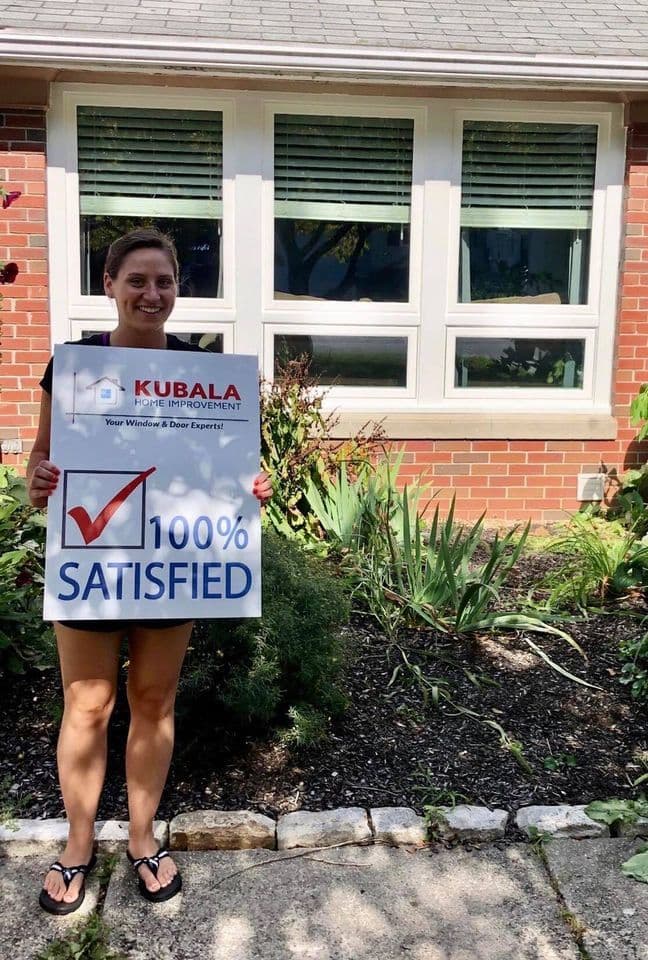 A homeowner holds a sign that reads "100% Satisfied" and stands in front of newly-replaced windows.