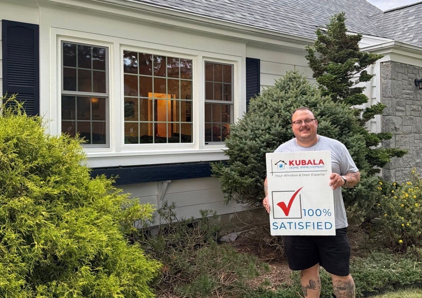Homeowner holds a sign that reads "100% Satisfied" and stand in front of a newly-installed window.