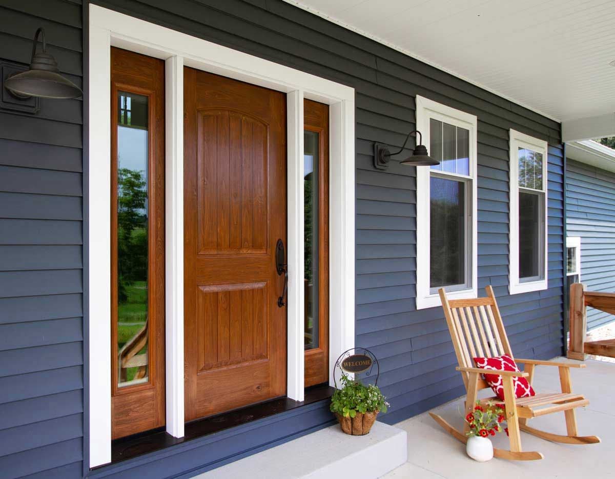A medium brown wood front door with narrow windows on either side on a dark navy blue house.